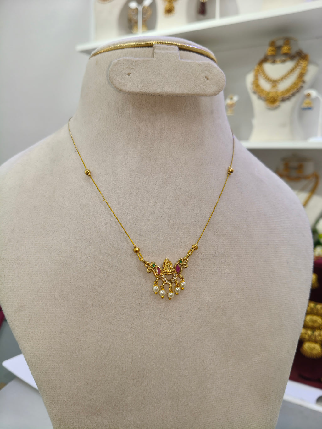 A gold necklace with a decorative pendant featuring small red and green stones and dangling pearl-like beads, displayed on a beige jewelry bust in a store setting.