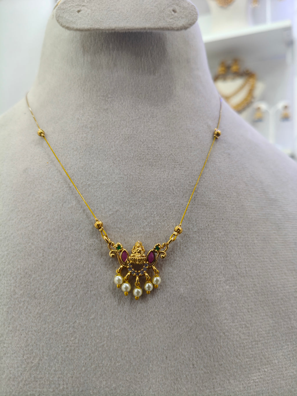Gold pendant necklace with a traditional Indian style ornament, featuring small white pearls and red gemstone accents, displayed on a gray jewelry bust.