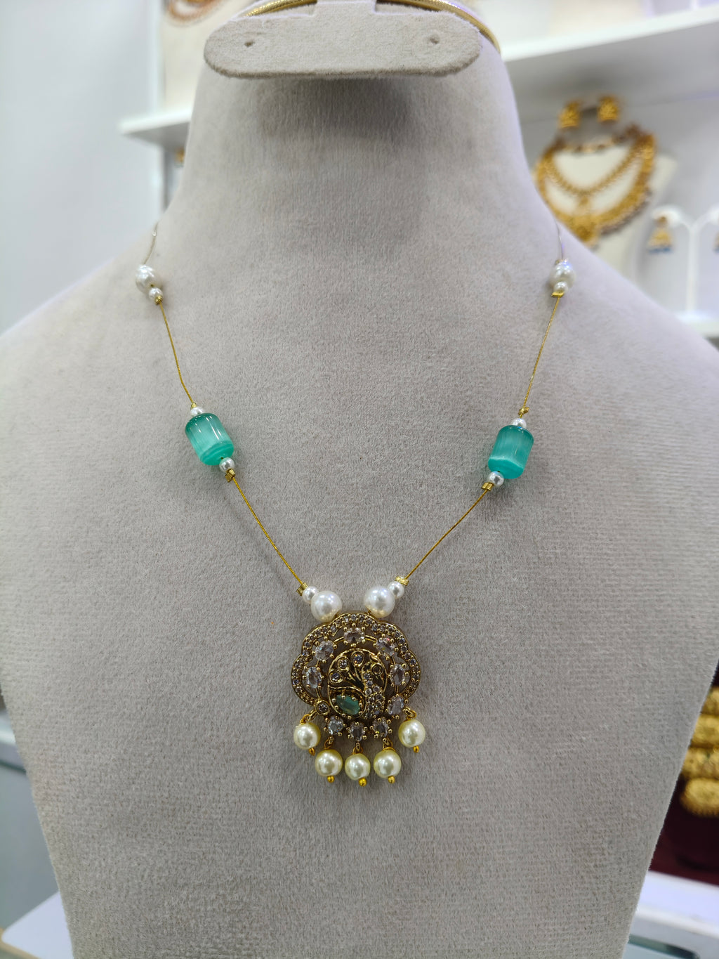 An ornate gold necklace displayed on a beige jewelry bust. The necklace features a detailed gold pendant with a central green stone, flanked by white pearl-like beads and translucent turquoise-green rectangular beads on a gold chain.