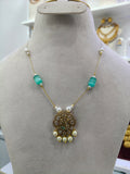 An ornate gold necklace displayed on a beige jewelry bust. The necklace features a detailed gold pendant with a central green stone, flanked by white pearl-like beads and translucent turquoise-green rectangular beads on a gold chain.