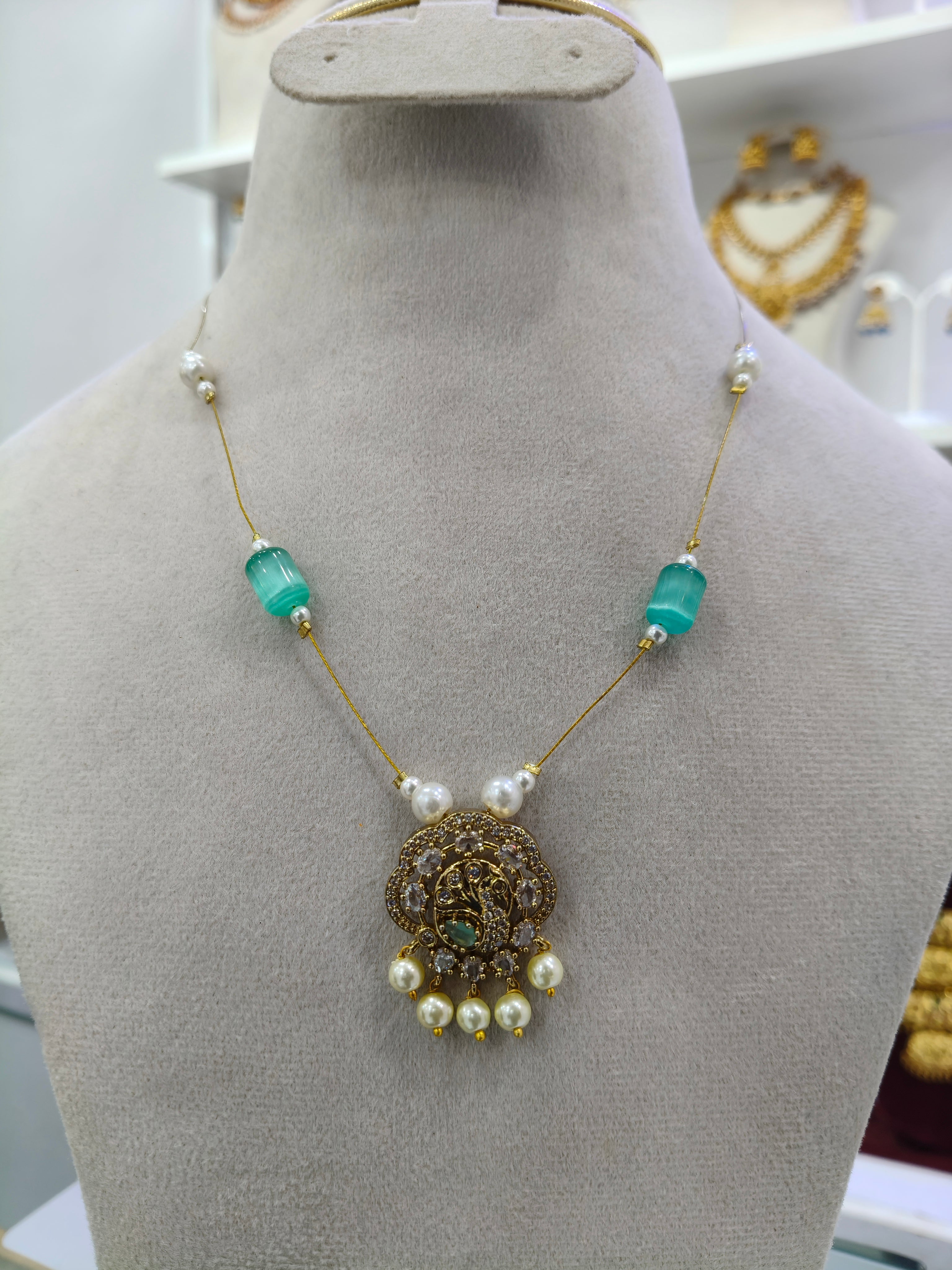 An ornate gold necklace displayed on a beige jewelry bust. The necklace features a detailed gold pendant with a central green stone, flanked by white pearl-like beads and translucent turquoise-green rectangular beads on a gold chain.