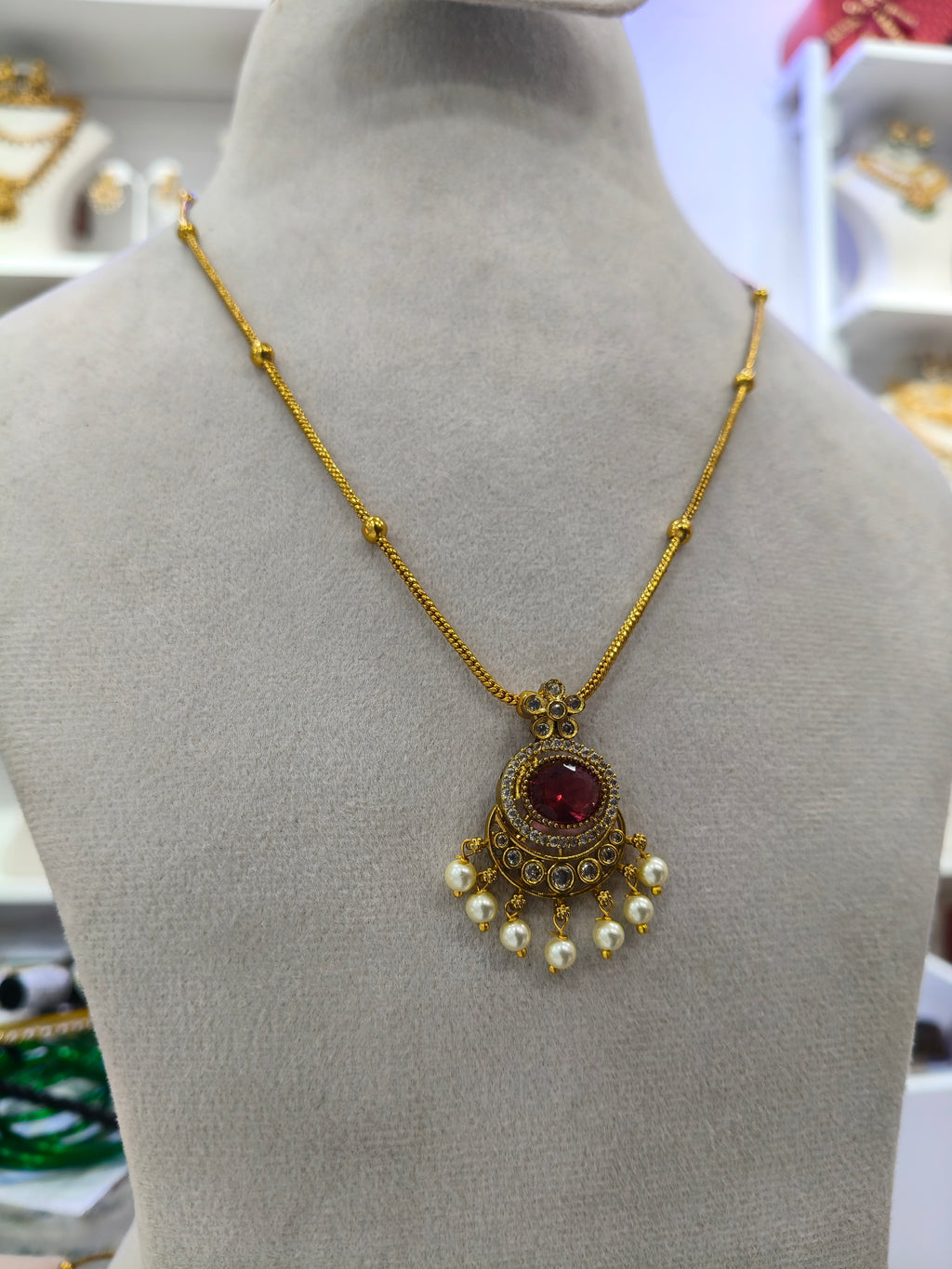 Gold chain necklace with a circular red gemstone pendant and dangling white pearls, displayed on a gray mannequin bust.