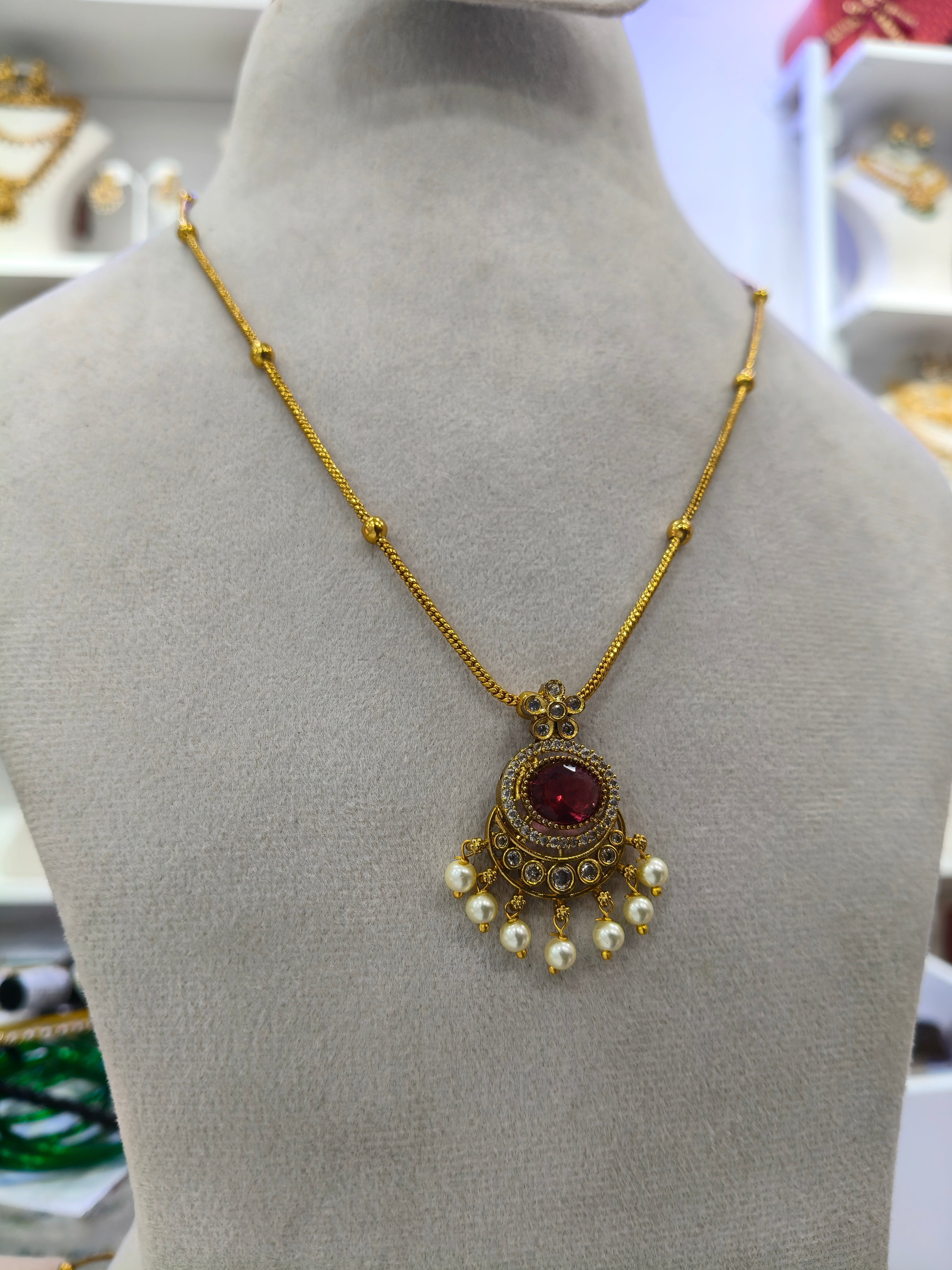 Gold chain necklace with a circular red gemstone pendant and dangling white pearls, displayed on a gray mannequin bust.